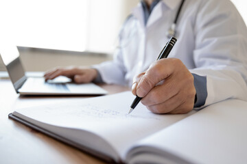 Fototapeta premium Crop close up of middle-aged male doctor write in medical journal fill patient information use laptop online. Mature man GP or therapist make notes work distant on computer in clinic or hospital.