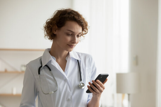 Happy Young Caucasian Female Nurse Or GP In White Uniform Look At Cellphone Screen Use Gadget In Hospital. Smiling Woman Doctor Consult Patient Or Talk With Clinic Client On Video Call On Smartphone.