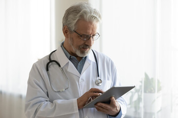 Mature Caucasian male doctor in white medical uniform use modern tablet in hospital or clinic. Middle-aged grey-haired man GP or therapist consult patient online on pad device. Healthcare concept. - obrazy, fototapety, plakaty