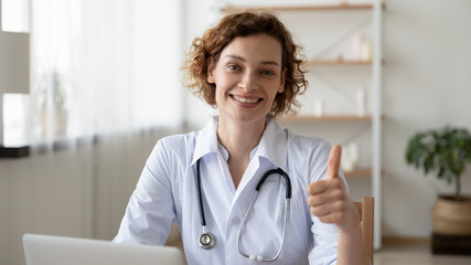 Portrait of smiling young Caucasian female doctor in white medical uniform show thumb up give...
