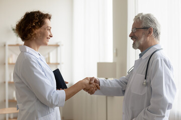 Smiling diverse doctors in white medical uniforms shake hands get acquainted in hospital. Happy...