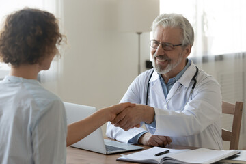 Obraz premium Smiling elderly male doctor shake hang greet get acquainted with female patient at meeting in clinic. Happy mature man GP handshake woman close health insurance deal at consultation in hospital.
