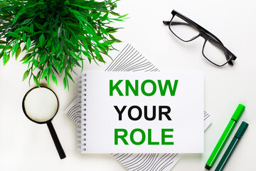 On a white background lies a notebook with words KNOW YOUR ROLE, glasses, a magnifying glass, green markers and a green plant