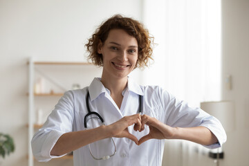 Portrait of happy young Caucasian female doctor in white medical uniform show heart love hand...