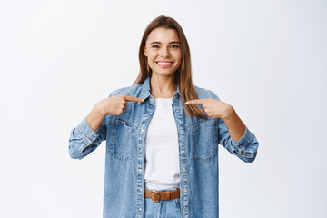 Smiling confident girl pointing at herself for self-promoting, self-appreciate her own achievement, boasting with accomplishments, standing against white background