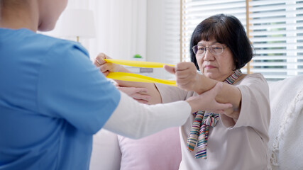 Young older senior asia citizen sit on sofa couch with in-home nursing care, assisted living, scrubs nurse doctor use resistance band exercise for senior patient in physiotherapy treatment at home.