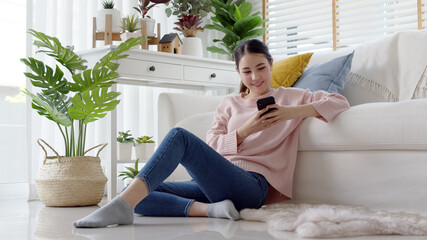 Young attractive beautiful casual asia female lady student look at cellphone play social media sit at sofa couch living room feel relax comfort carefree with green houseplant at cozy home apartment.