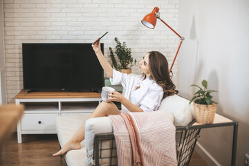 Young woman makes selfie sitting on couch at home. Young person drinks coffe in big mug and use smartphone