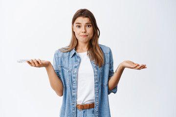 Nervous young woman shrugging with hands spread sideways, holding phone and frowning troubled, having problem on smartphone, standing over white background