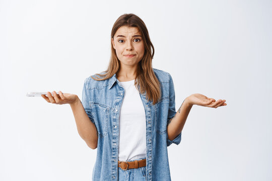 Nervous Young Woman Shrugging With Hands Spread Sideways, Holding Phone And Frowning Troubled, Having Problem On Smartphone, Standing Over White Background