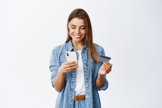 Online shopping. Portrait of smiling blond woman paying with plastic credit card on smartphone app, standing with mobile phone and bank card against white background