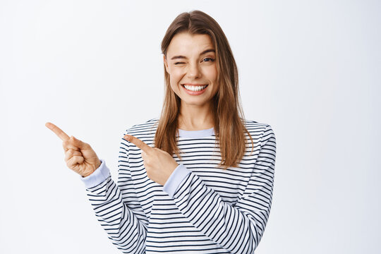 Cheerful Smiling Woman Inviting To Check Out Promo, Pointing Fingers Left And Looking Happy, Winking At Camera To Motivate You, White Background