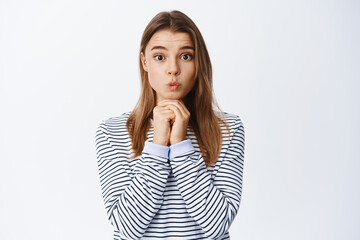 Cute and silly blond girl looking with temptation, holding hands above chin, pucker lips and...
