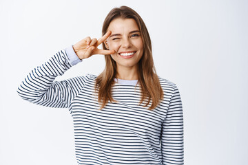 Portrait of positive young woman winking and showing v-sign over eye, smiling with white perfect...