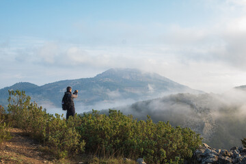 Young hiker observing the landscape and taking photos from a mountain, in a foggy morning.