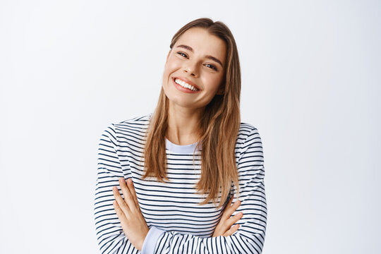 Image of beautiful blond girl with white smile, tilt head and grinning happy at camera, cross arms on chest, standing in casual clothes against white background