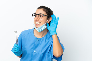 Young woman dentist holding tools isolated on white background listening to something by putting hand on the ear