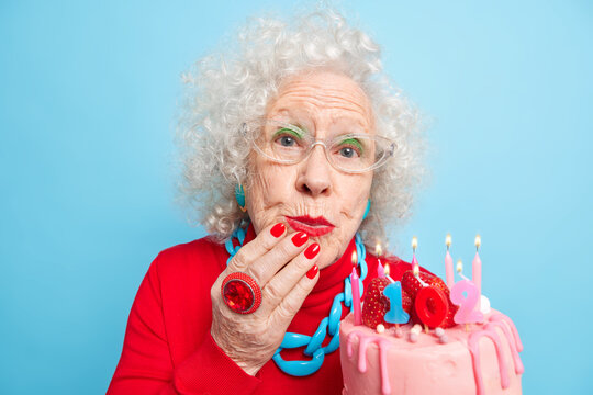 Portrait of good looking beautiful woman wears jewelry and festive red clothes holds cake with candles celebrates 102nd birthday being on party for elderly people isolated over blue background