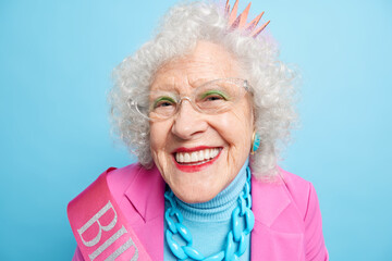 Close up shot of cheerful elderly birthday woman looks happily at camera wears princess crown spectacles stylish clothes celebrates her 80th anniversary isolated over blue background. Aging concept