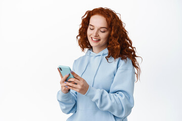 Fototapeta premium Beautiful redhead teen girl using mobile phone chat app, looking at smartphone, reading mobile screen and smiling, standing against white background