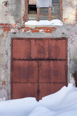 An old rusty snow-covered gate in the wall of an abandoned industrial building. The entrance is closed and blocked.