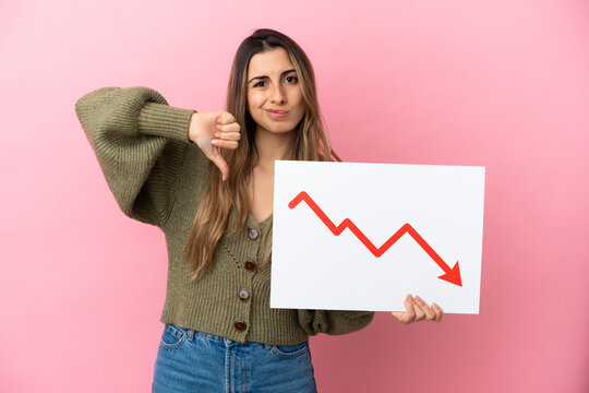 Young caucasian woman isolated on pink background holding a sign with a decreasing statistics arrow symbol and doing bad signal