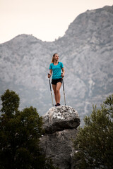 Obraz premium view of young woman traveler with trekking poles standing on large stone