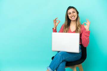 Naklejka premium Young caucasian woman sitting on a chair with her pc isolated on blue background showing an ok sign with fingers