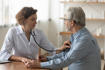 Caring Caucasian female doctor use phonendoscope examine mature male patient heart rate at...