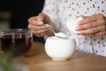 Just one teaspoon is enough. Close up cropped shot of young lady brew strong black herbal flower tea add heaped spoon of granulated sugar for a cup. Focus on transparent teacup with hot drink and bowl