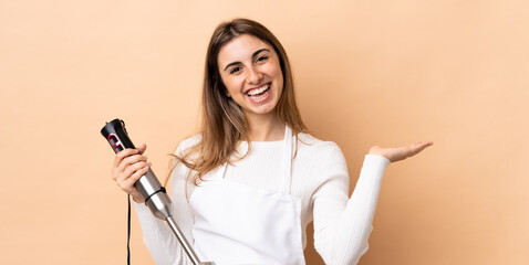 Woman using hand blender over isolated background holding copyspace imaginary on the palm to insert an ad