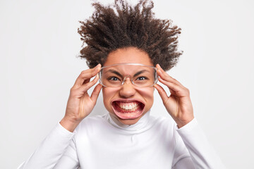 Close up shot of dark skinned curly woman keeps hands on spectacles cleches teeth feels outraged and crazy dressed in casual clothes isolated over white background. Negative emotions concept