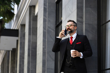 Handsome Caucasian business man using mobile phone and holding take away coffee cup outside apartment in the morning before going to work