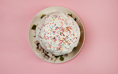 Easter composition. A glazed Easter cake decorated with sugar sprinkles and apricot flowers on a white plate stands on a pastel pink background. Happy Easter Holidays. Top view.