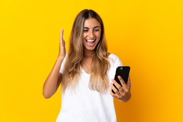 Fototapeta premium Young hispanic woman over isolated yellow background looking at the camera while using the mobile with surprised expression
