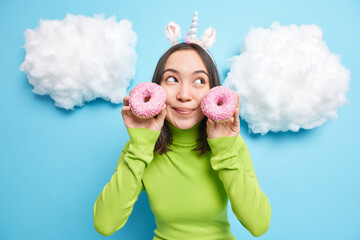Dreamy brunette Asian woman holds two glazed donuts near face has sweet tooth concentrated aside imagines how she eats this tasty dessert wears unicorn headband green turtleneck stands indoor