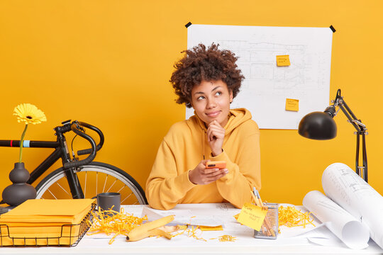 Dreamy pleased Afro American woman holds modern cellular makes building blueprints works on sketches wears sweatshirt has mess on office desk isolated over yellow background. Talented architect