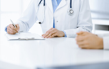 Fototapeta premium Unknown woman-doctor and female patient sitting and talking at medical examination in clinic, close-up. Therapist wearing blue blouse is filling up medication history record. Medicine concept