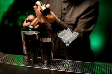 view on martini glass and shaker cups into which bartender pours drink from jigger