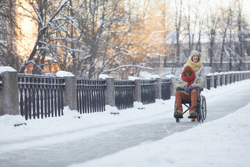 Wide angle view at African American man using wheelchair having fun outdoors in winter with smiling young woman assisting, copy space
