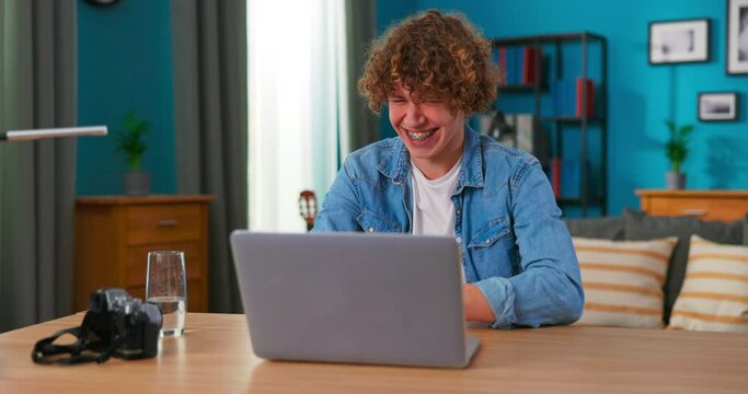 Successful teenage boy sits at table uses laptop read e-mails online home office, gets good news, positive exam results, new opportunity, feels joy triomphe, celebrates victory, happy dancing