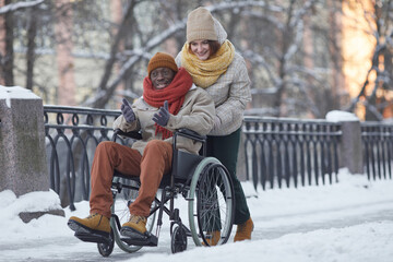 Full length portrait of African American man using wheelchair having fun outdoors in winter with smiling young woman assisting, copy space