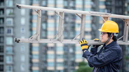 Asian maintenance worker man with protective suit and safety helmet carrying aluminium step ladder at construction site. Civil engineering, Architecture builder and building service concepts