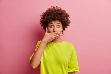 Fototapeta na wymiar Horizontal shot of surprised Afro American woman tries to be speechless covers mouth with palm stares shocked at camera hears gossips dressed in casual t shirt isolated over pink background.