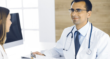 Doctor and patient discussing medical exam results while sitting at the desk in clinic. Male physician using clipboard for filling up medication history record of young woman. Data in medicine