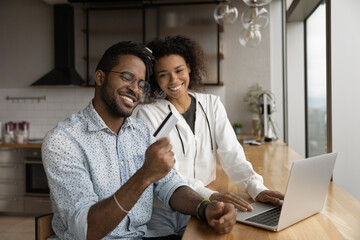 Close up happy African American couple paying online by credit card, using laptop, smiling wife and...