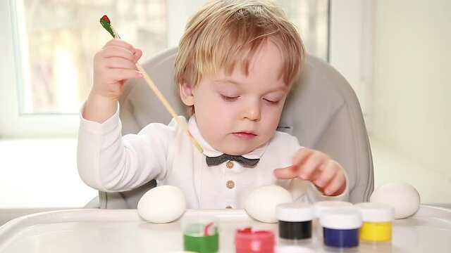 The blonde toddler uses colored paint from a jar and a paintbrush to paint Easter eggs. A carefree and happy childhood. Selective Focus