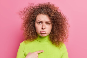 Offended insulted pretty teenage girl with curly bushy hair points at herself purses lower lip has displeased expression wears green turtleneck isolated over pink background asks do you blame me