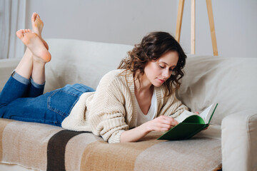 Keen young woman reading in comforts of her apartment. She's lying on a couch on her stomach, crossing her bare feet.