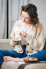 Relaxed young woman sewing in comforts of her apartment. She is sitting on a couch, embroidering a...